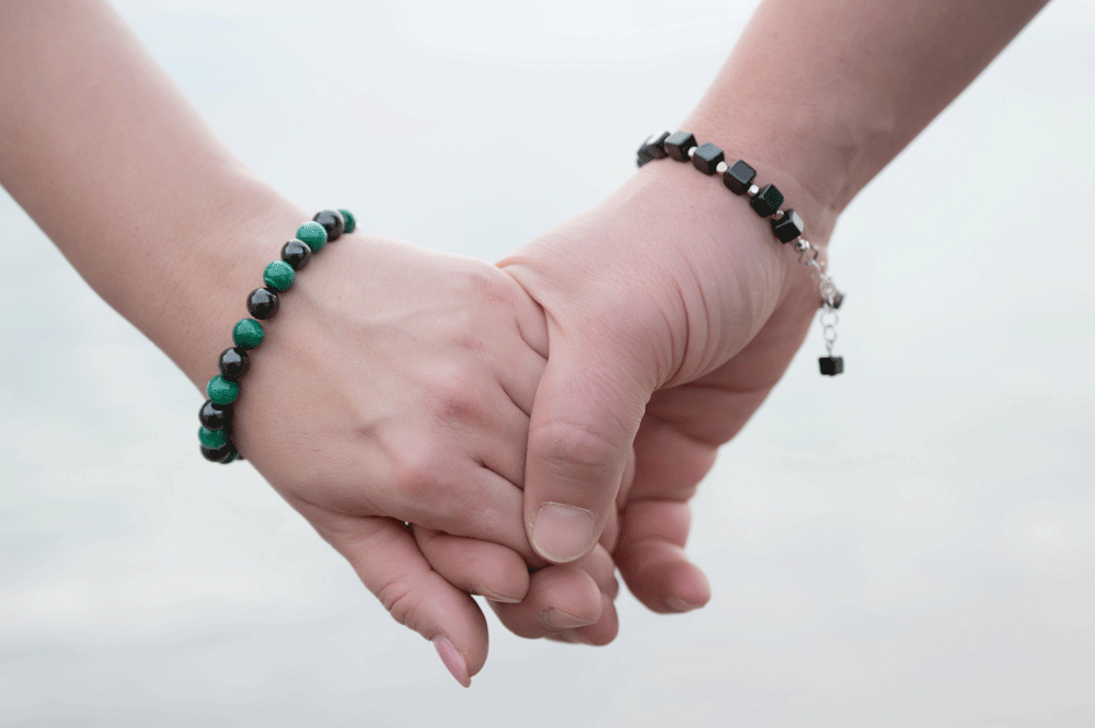 Zwei Hände, eine weibliche und eine männliche, die sich halten, jeweils mit eleganten Schungit-Armbändern am Handgelenk